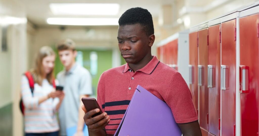 Teenage boy looking at phone with other teenagers in the background watching him. Learn how cyberbullying prevent can help a teen with autism.