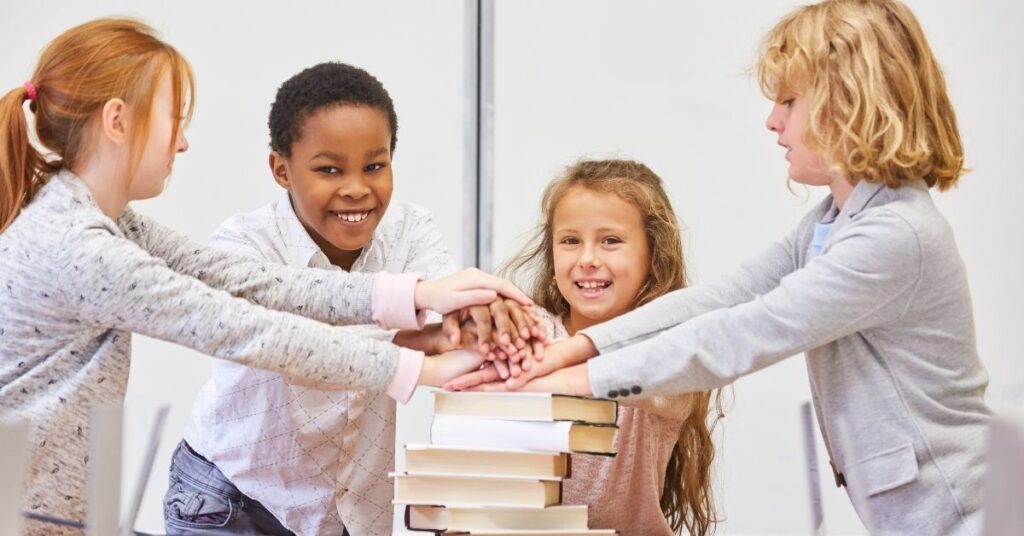 Four children holding their hands on top of each other on a stack of books. Social skills groups can help children with autism.