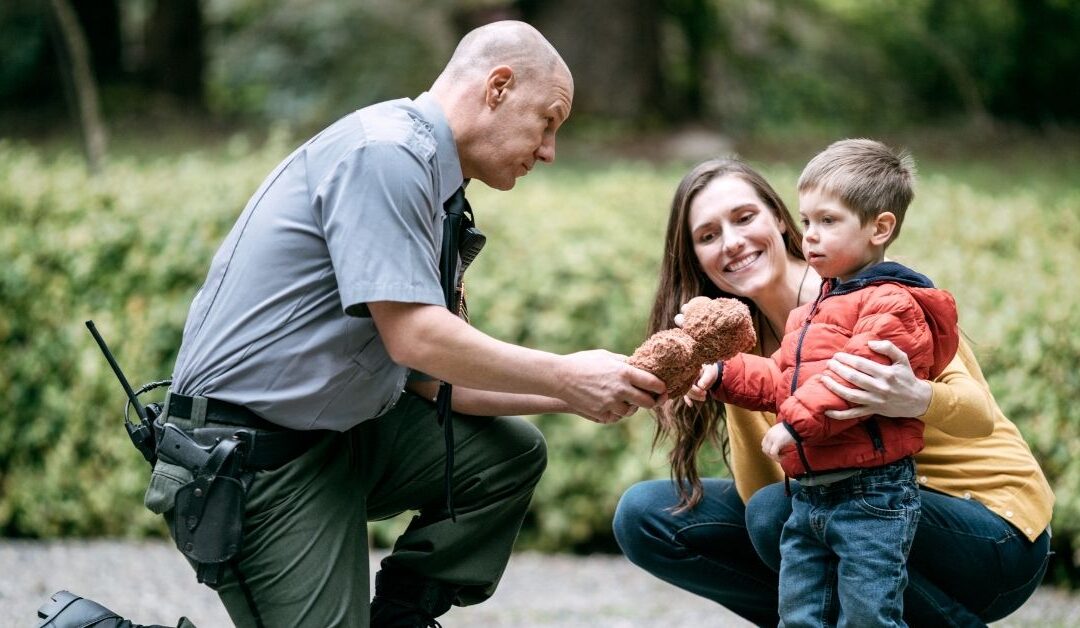 Police officer giving teddy bear to boy with mom nearby.