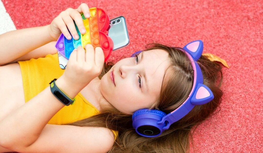Girl wearing headphones, lying on the ground with phone and sensory toy in hand