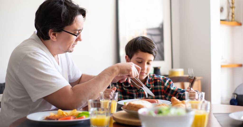 Parent sitting with boy at table and cutting up a hot dog on his plate. How to help your child with autism and ADHD overcome picky eating.
