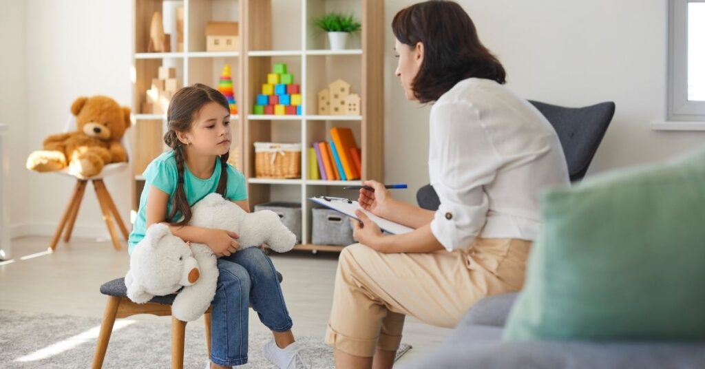 Girl sitting in a chair and holding a large stuffed bear, talking to a therapist across from her. Reasons to apply for the Medicaid Waiver for your child with autism.
