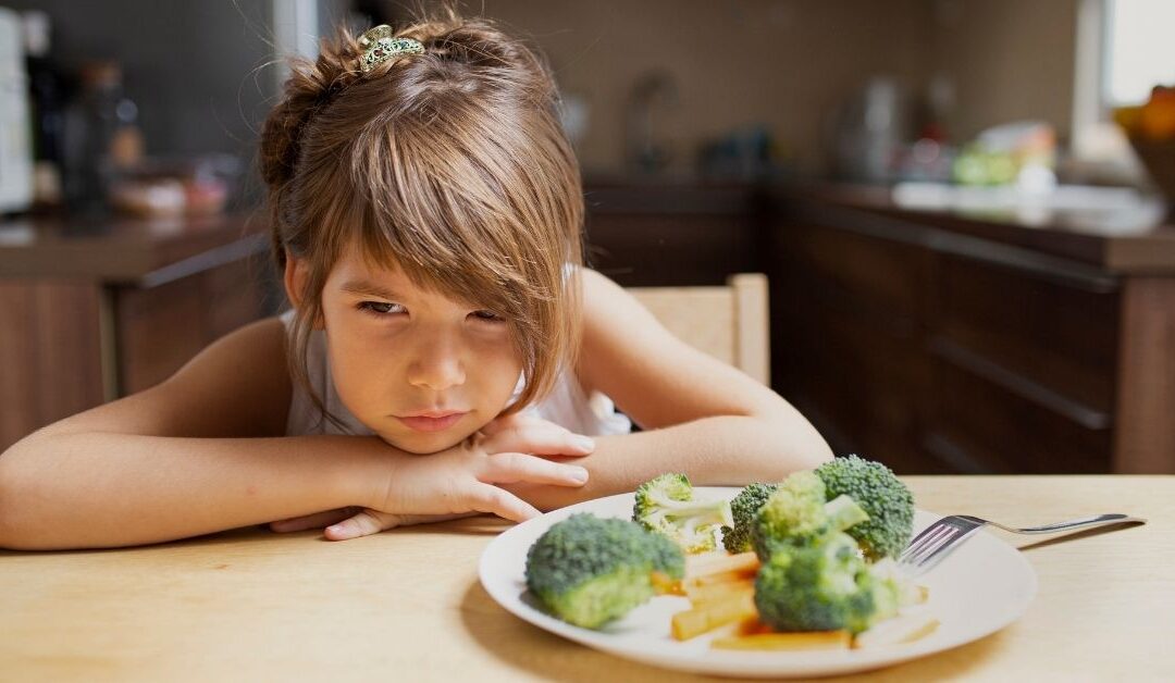 Girl with head on the table looking defiant with a plate of vegetable sitting next to her.