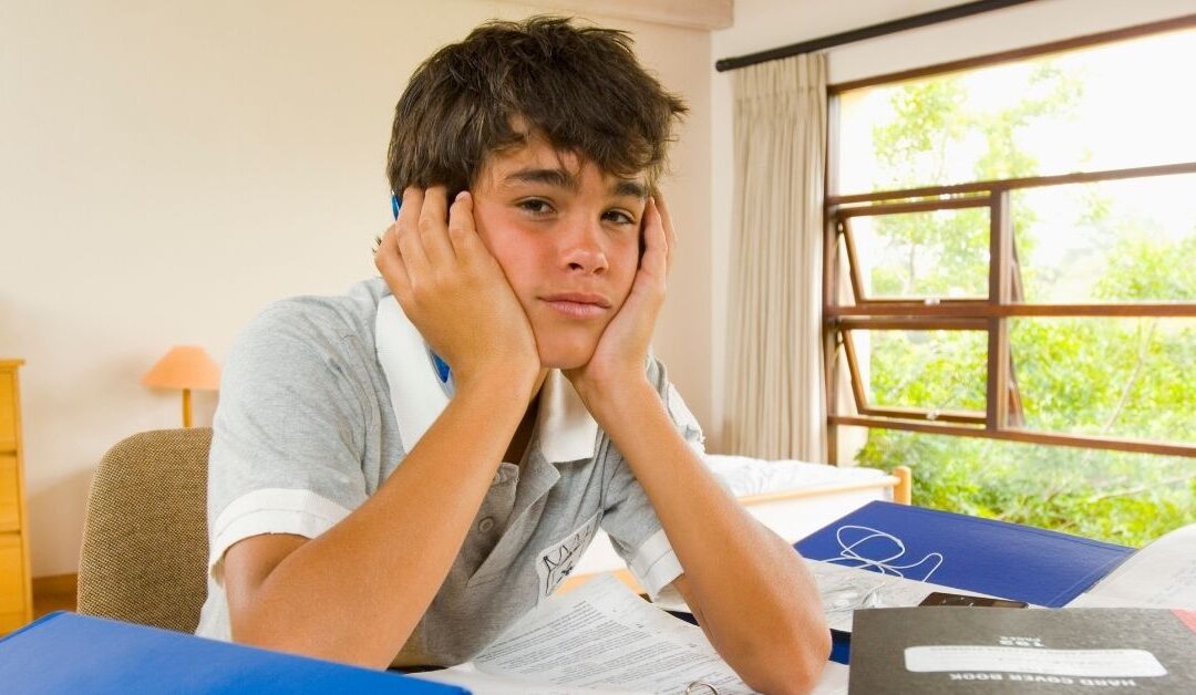 Teenage boy sitting among books with head in hands frustrated.