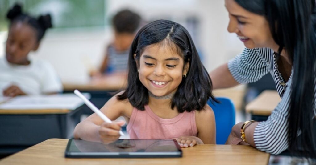 Girl working on tablet with teacher sitting next to her. How can technology help your autistic child?