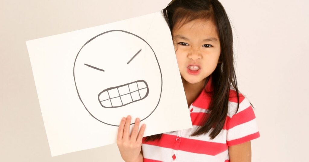 Girl with angry face holding a drawing of an angry face. How can you teach your autistic child how to gain emotional control?