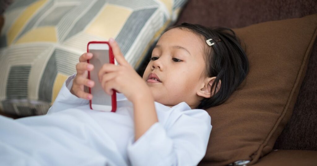 Little girl lying on couch looking at a smartphone. What are the pros and cons of screen time for a child with autism and ADHD?