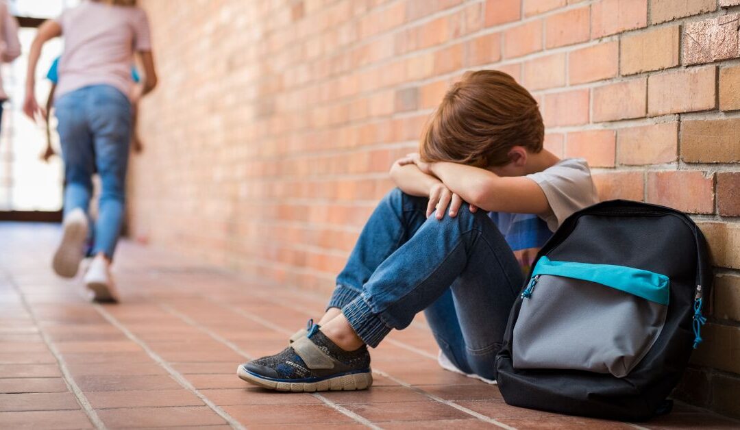 Boy sitting on floor against school wall with head in his arms, seeming unhappy