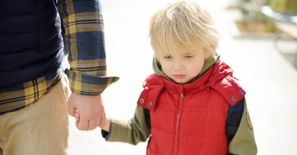 Boy holding his father's hand, walking and looking unhappy. How can you help your child with autism overcome school avoidance or refusal?