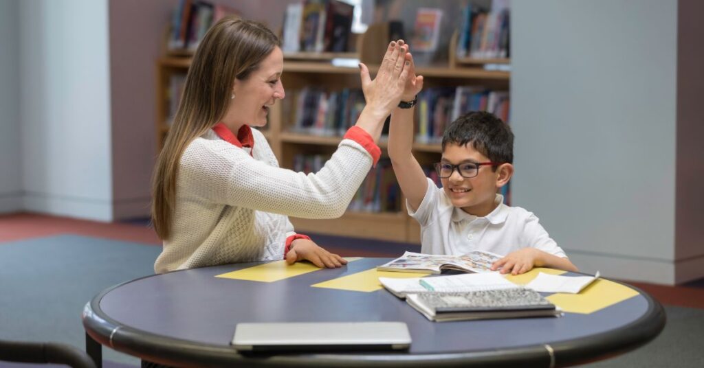 Tutor giving boy a high five as they sit at a table with books. How to determine if your autistic child needs a tutor.