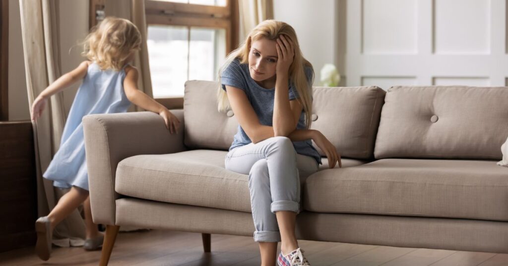 Mom sitting on couch with head in her hands looking stressed while little girl runs around the couch