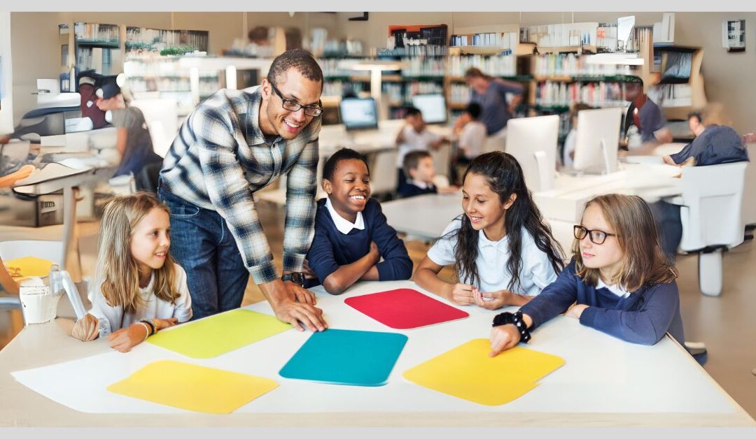 Teacher standing at table with students that have colorful sheets of paper in front of them on a table