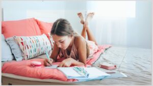 Early teen girl lying on bed writing on tablet. How to support your autistic teenager through their adolescent years.