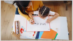 Mom helping her daughter learn how to write on a large paper on a small table while sitting on the floor. Would homeschooling be a good option for your autistic child?