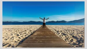 Teen boy standing on boardwalk on a beach going out to lake with his arms wide open